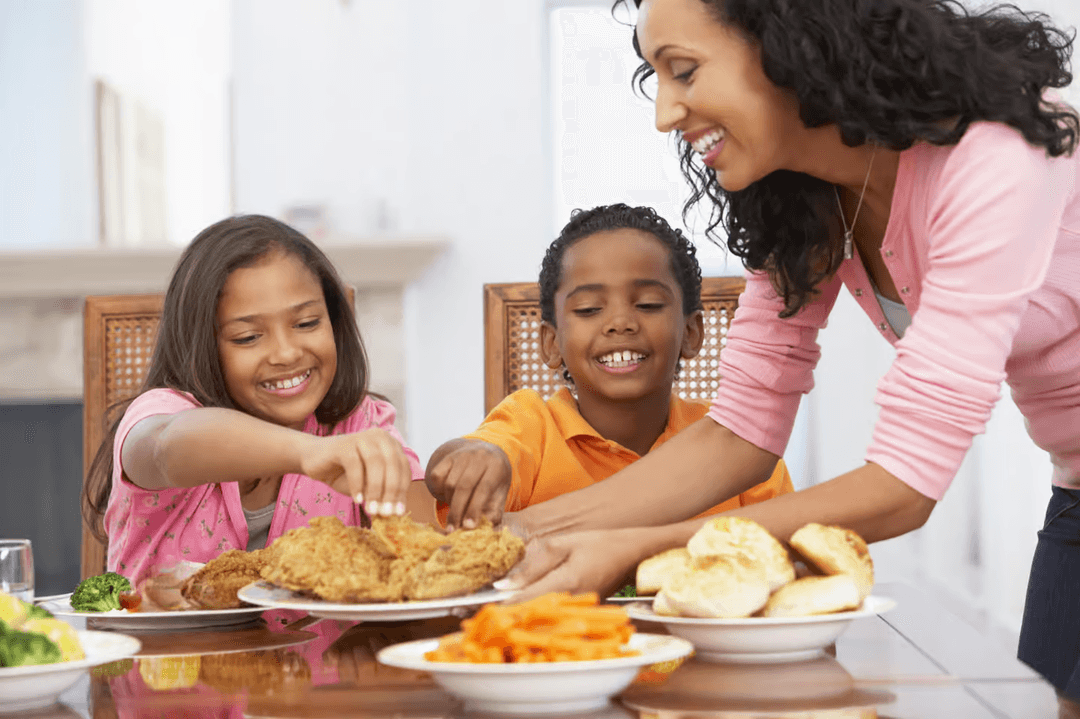 A mother and two children smiling and reaching for fried chicken and sides at a family dinner table